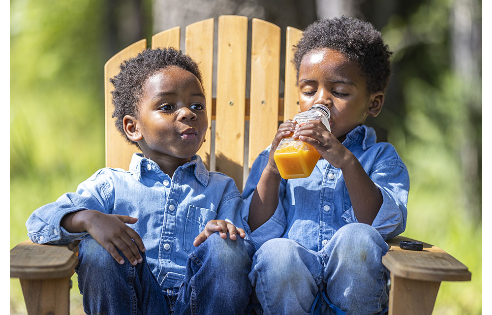Twin boys drinking orange juice while sitting together on a wooden chair.